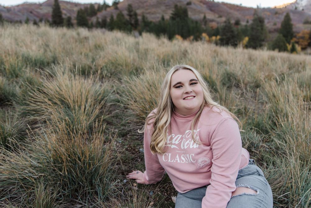 Person wearing pink sweatshirt sits in tall grass with mountains in background.