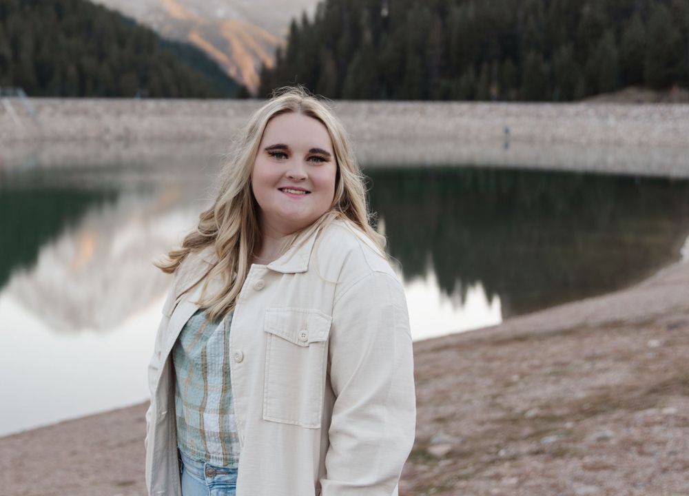 Person in cream jacket and plaid top poses by mountain lake at sunset.