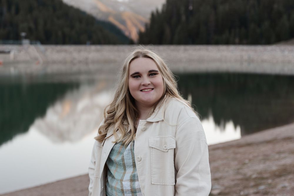 Portrait by serene mountain lake with person wearing light jacket and plaid shirt.