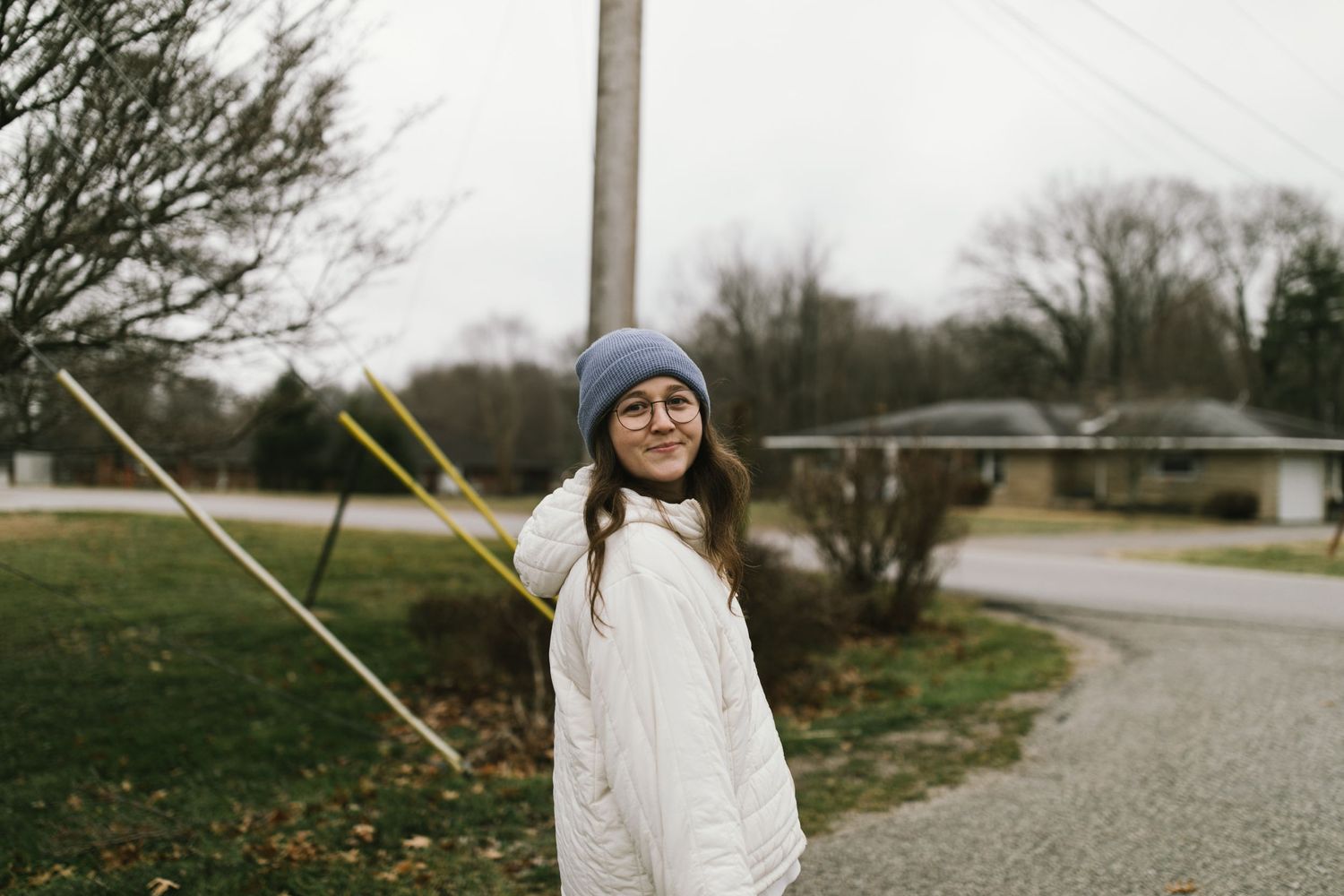 Person in white coat and beanie poses on rural road with bare trees in background.