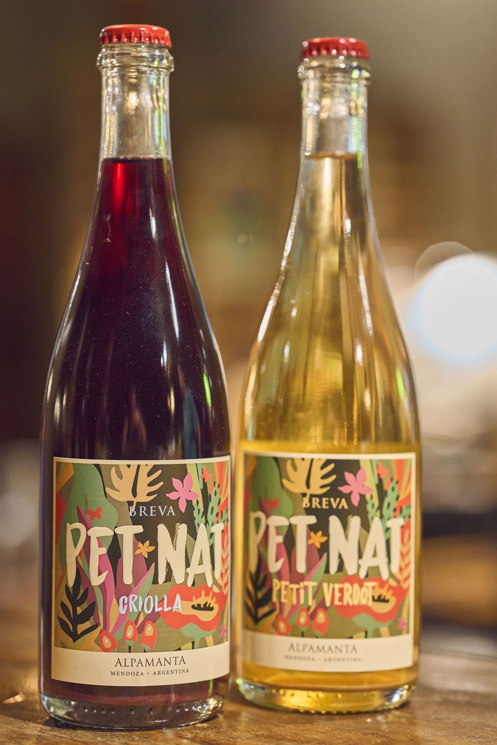 Two bottles of Pet-Nat wine from Alfamanta, one red and one light-colored, displayed on a wooden surface.