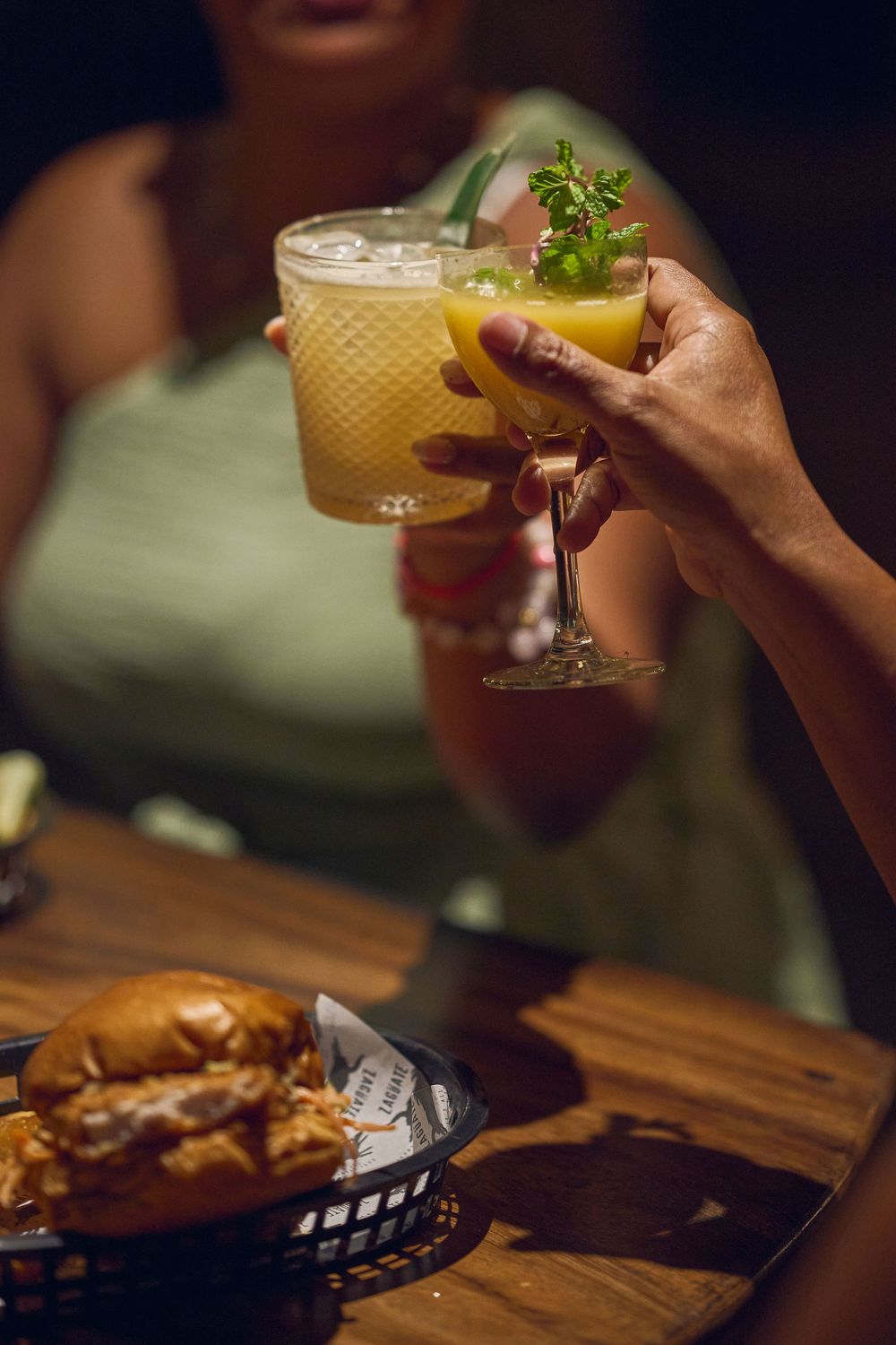Two hands toast cocktails garnished with herbs over a wooden table with a burger in a basket.