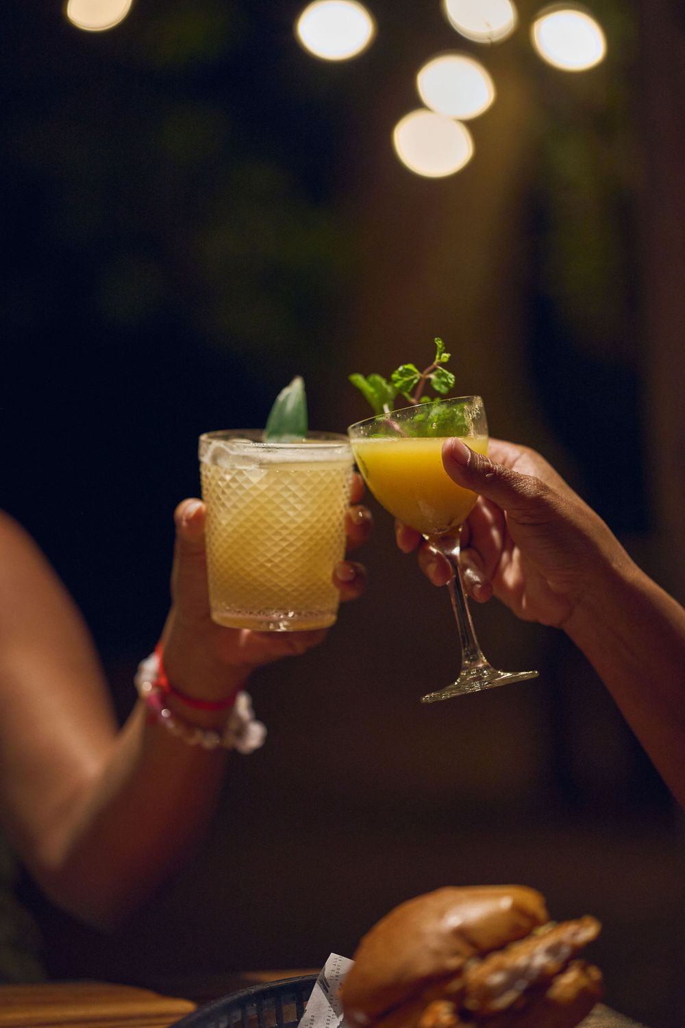Two cocktail glasses clink together against a backdrop of bokeh lights and a burger on a table.