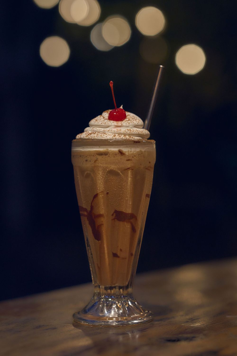 Chocolate milkshake topped with whipped cream and cherry served in a tall glass with bokeh background.