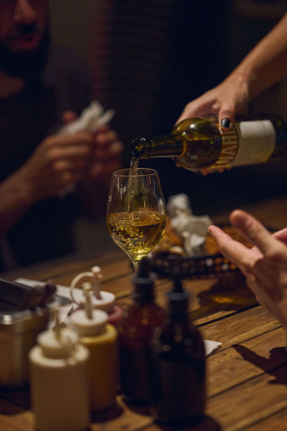 Multiple hands pour and serve golden-colored beverages on a rustic wooden table in a dimly lit setting.