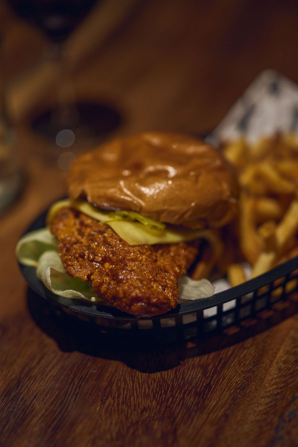 A crispy fried chicken sandwich with pickles served in a black plastic basket on a wooden table.