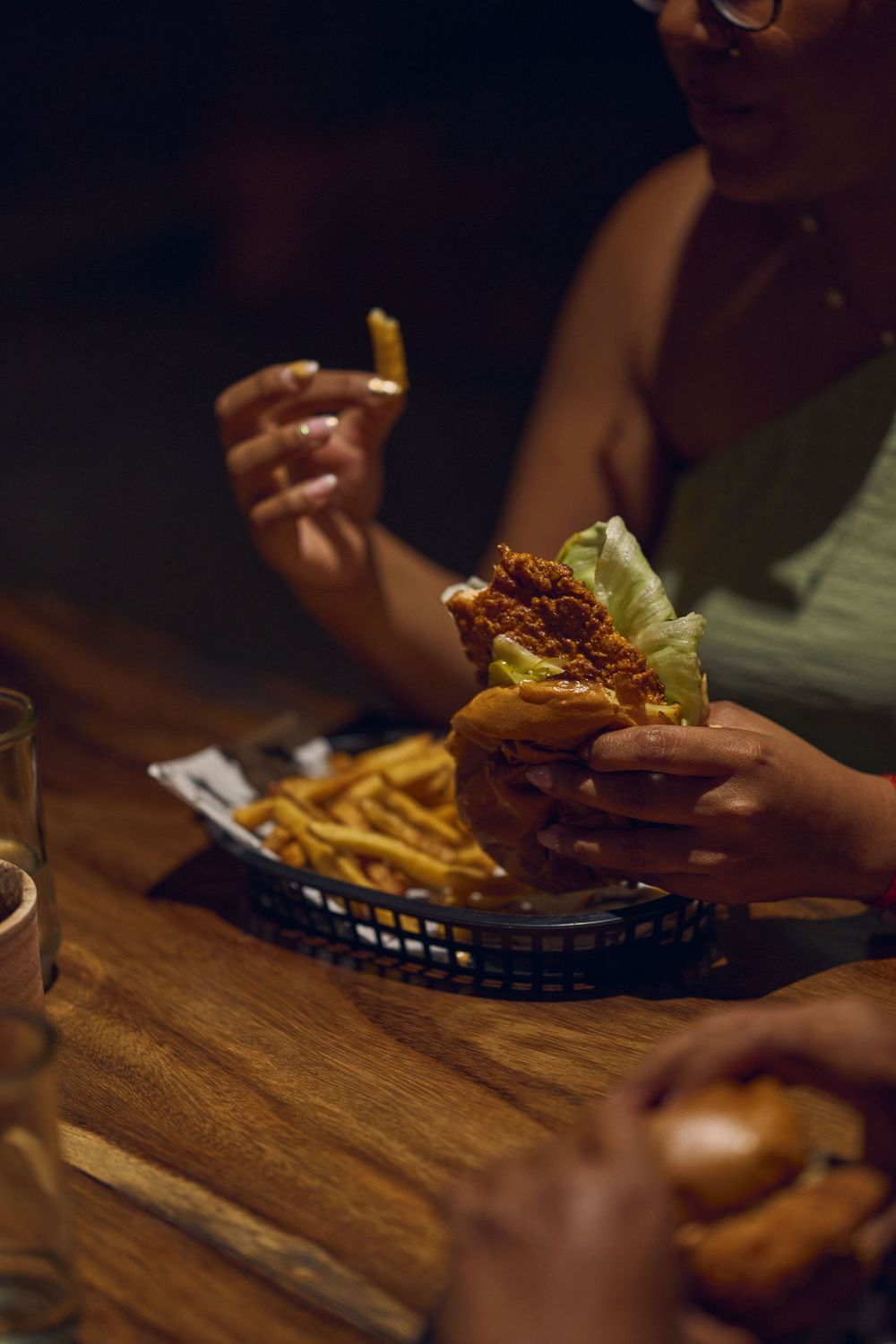Someone holds a juicy burger and french fries in a basket at a dimly lit restaurant with wooden table.