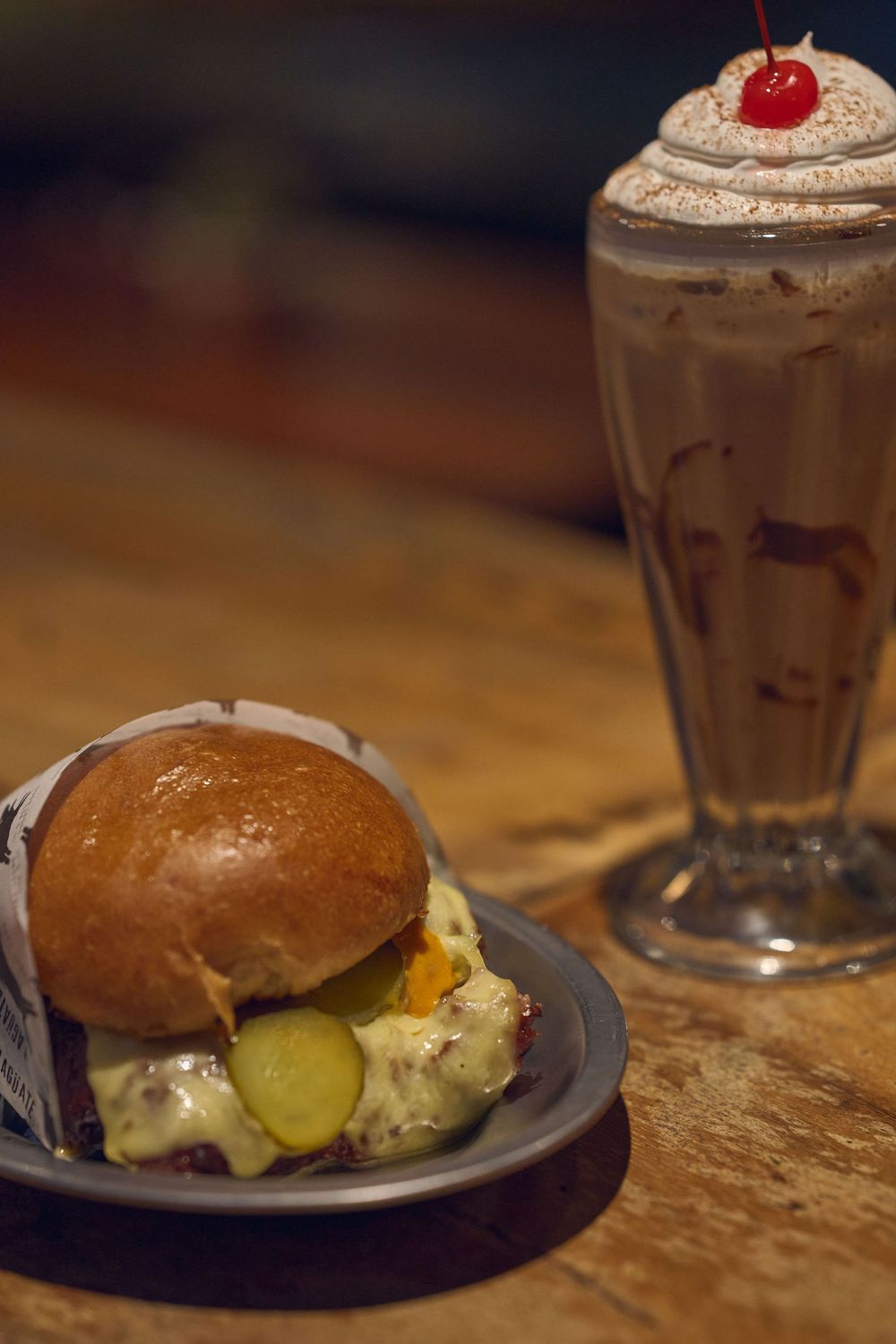 A gourmet burger with pickles served on a plate next to a tall chocolate milkshake topped with whipped cream and a cherry.