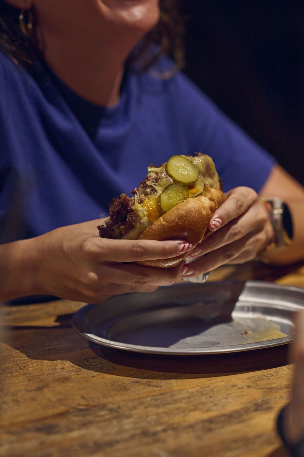 Close-up of hands holding a burger with pickles on a wooden table with plate in dark ambiance.