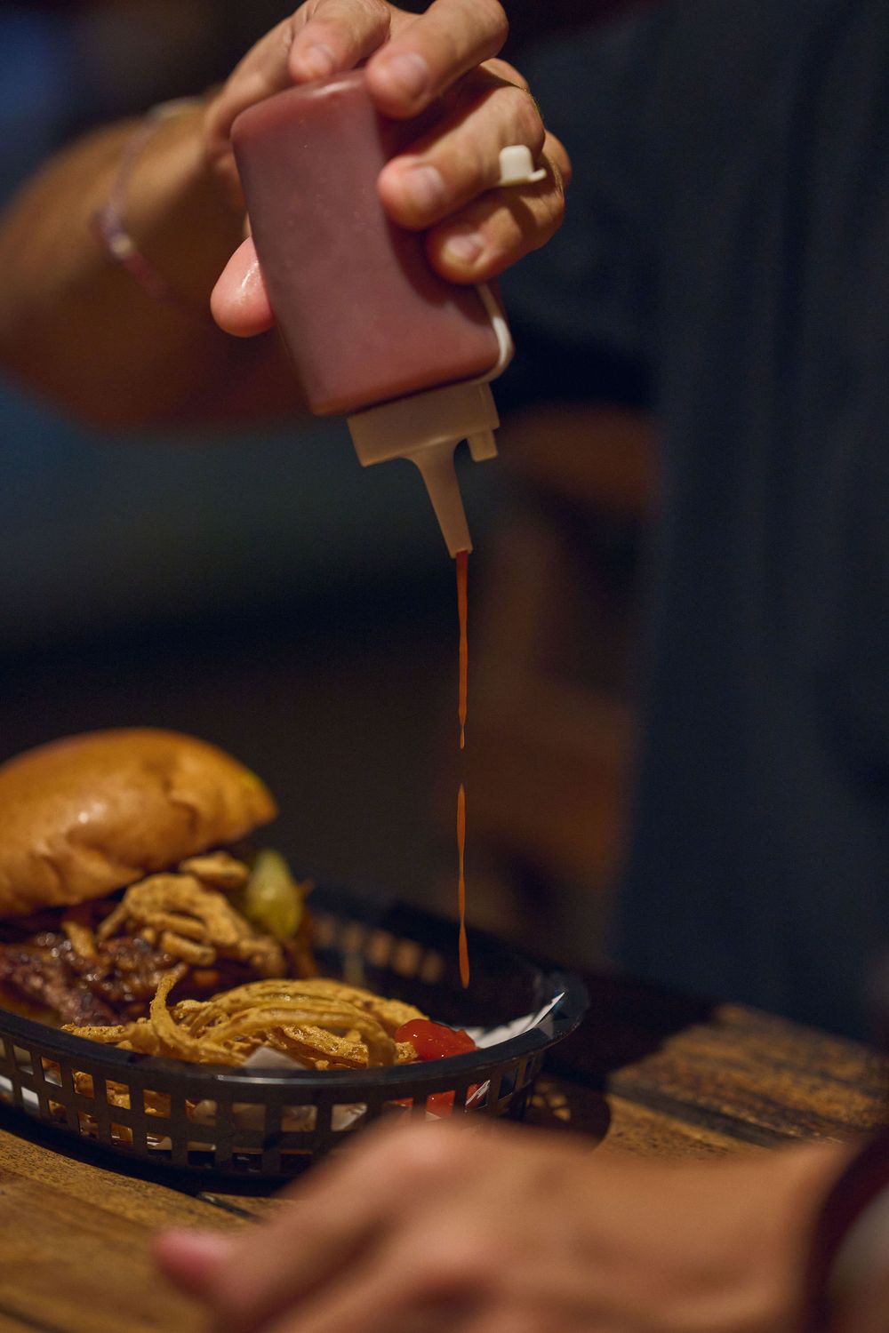 Red sauce being squeezed from bottle onto burger in basket on rustic wooden surface.