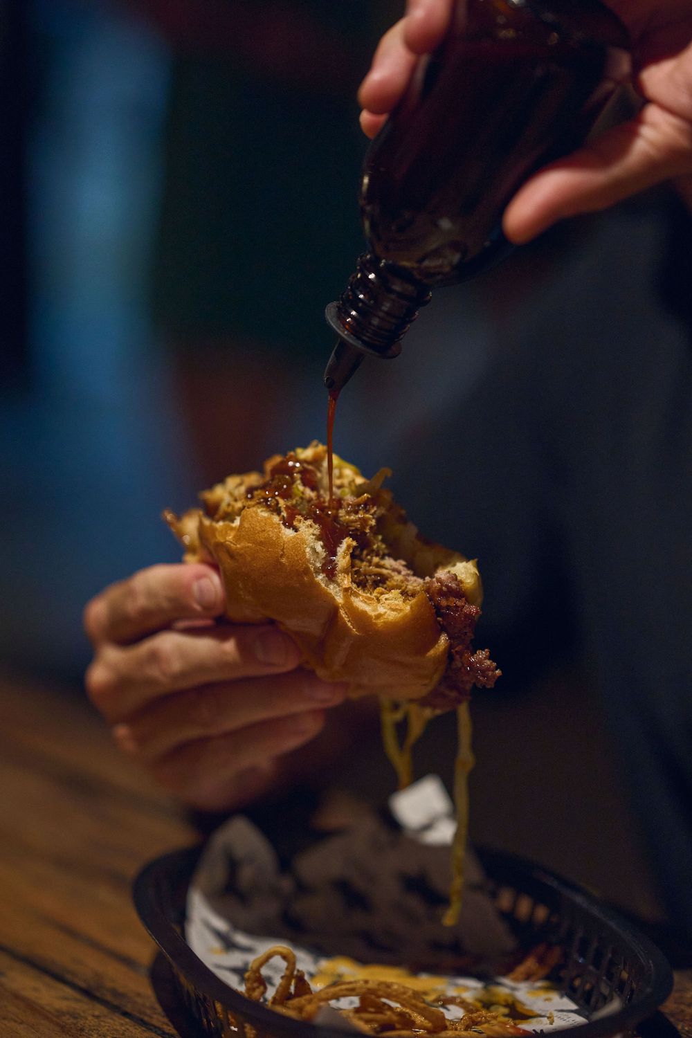 A hand holds open a juicy cheeseburger with melted cheese and toppings dripping onto a wooden surface.