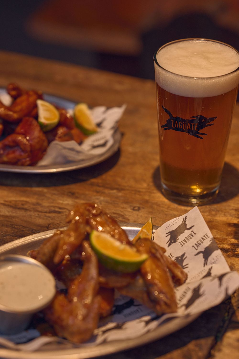 Plates of golden chicken wings served with beer and lemon slices on a rustic wooden table at a pub or restaurant.