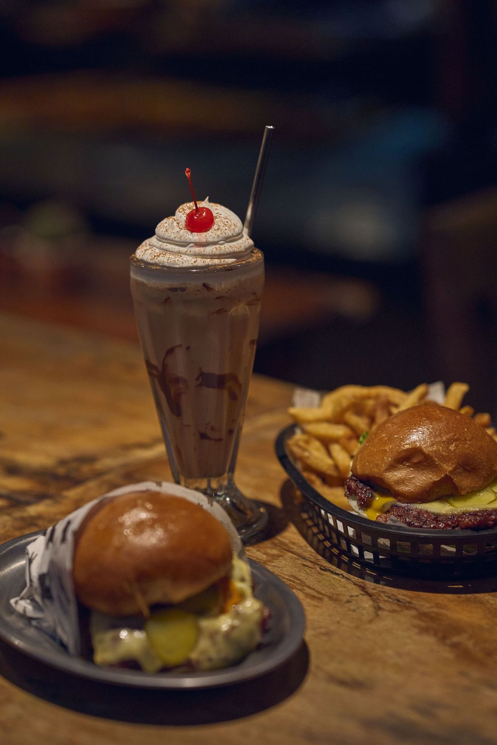Close-up shots of various decadent burgers and a chocolate milkshake served on rustic wooden tables at a restaurant.