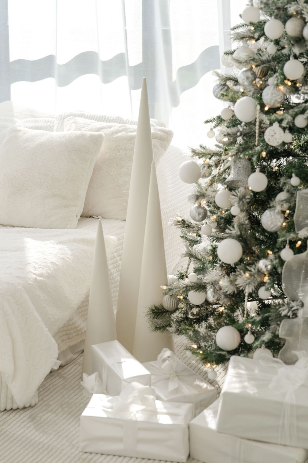 White and silver Christmas tree decorated with ornaments stands elegantly beside plush white cushions and wrapped presents.