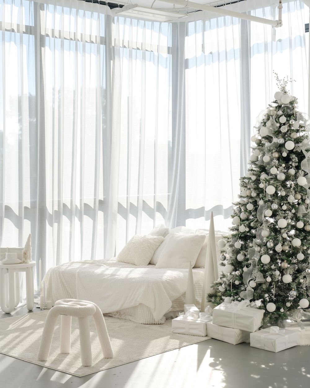 Bright sunlit room with white curtains, cozy white sofa and an elegantly decorated Christmas tree with presents.