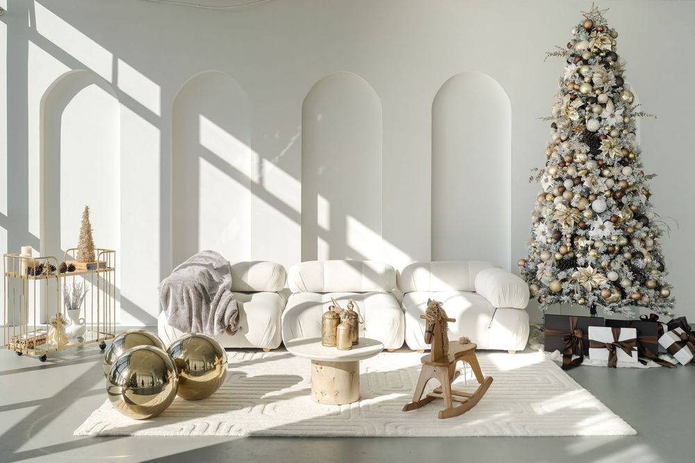 Minimalist white room with Christmas tree, rocking horse, and large gold ornaments in natural sunlight.
