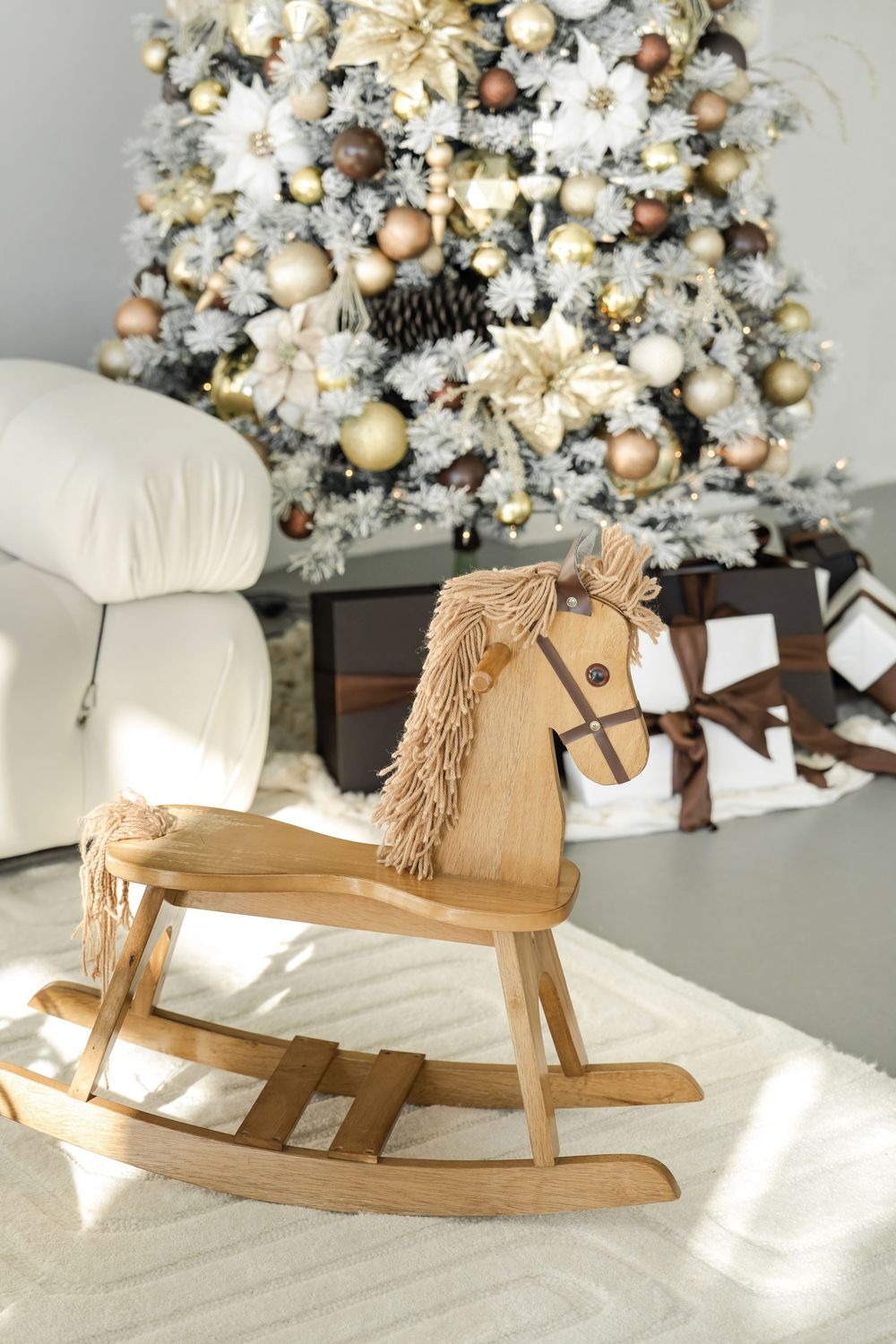 Wooden rocking horse sits in front of an elegantly decorated white and gold Christmas tree in a cozy living room.