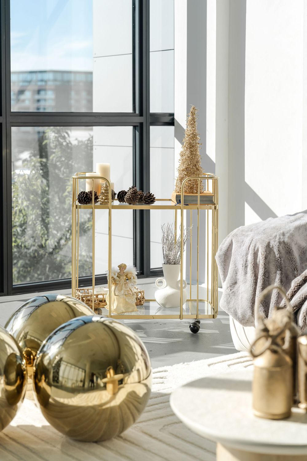 Gold Christmas decorations and ornaments displayed on a modern metal shelf near a sunny window.