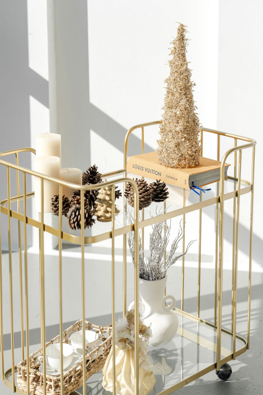 Modern gold serving tray decorated with pine cones and small gold glittery Christmas trees on white surface with dramatic shadows.