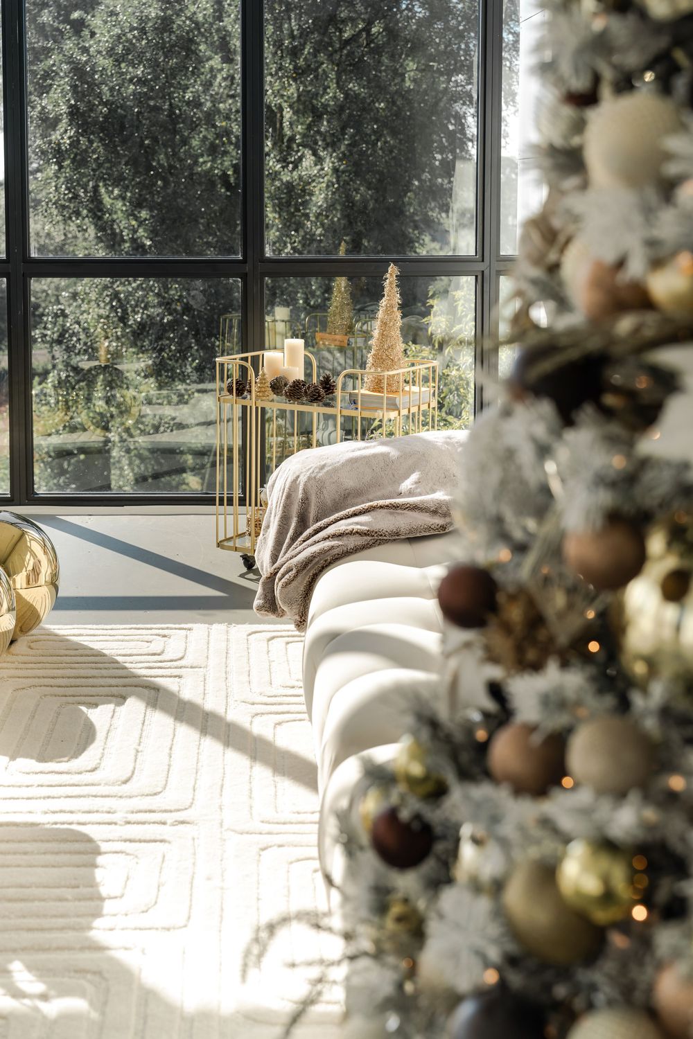 Cozy living room with a decorated Christmas tree, sunlit windows and gold holiday decor on a white accent table.