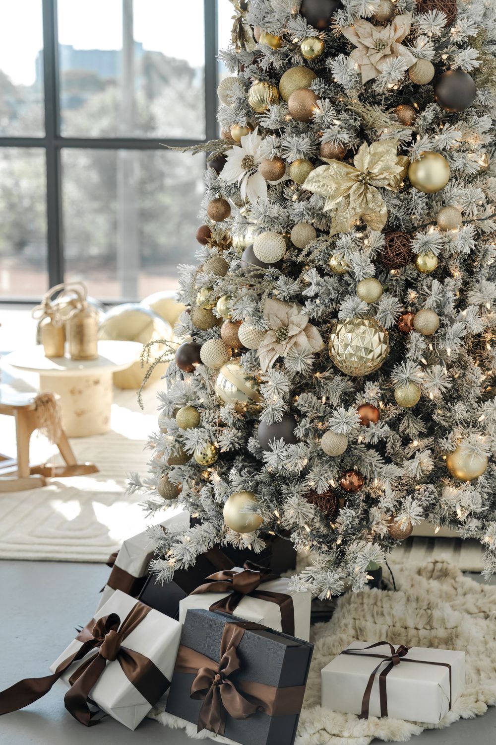 Elegantly decorated white flocked Christmas tree with gold and cream ornaments next to wrapped presents by a window.