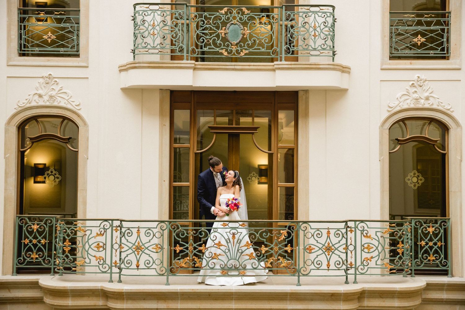 A couple poses on an ornate balcony with elegant wrought iron railings and arched entryways at a luxurious venue.