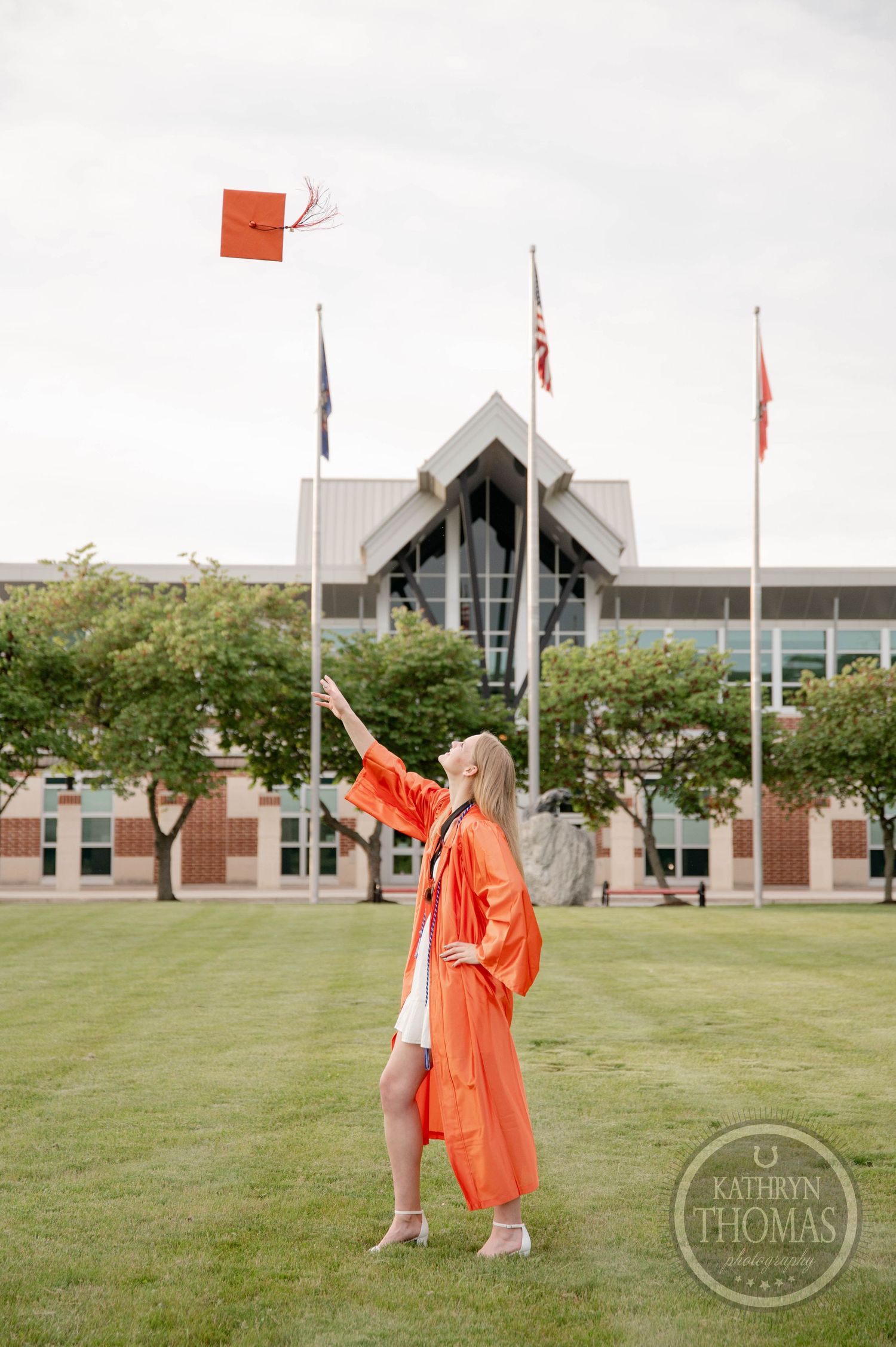 Graduate in orange cap and gown throws their graduation cap into the air on a sunny day in front of a campus building.