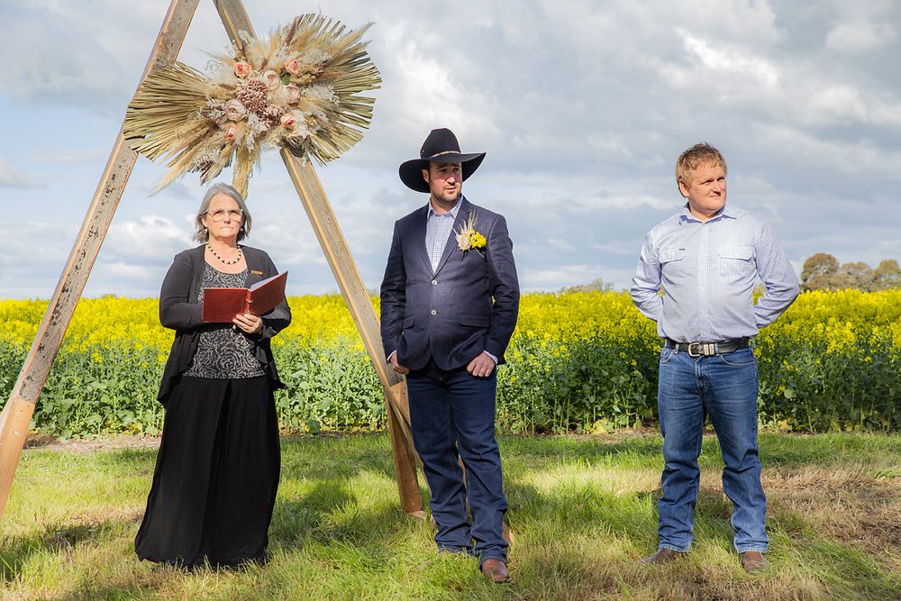 Three people stand in a field of yellow wildflowers with a decorative sunburst on a wooden frame behind them.