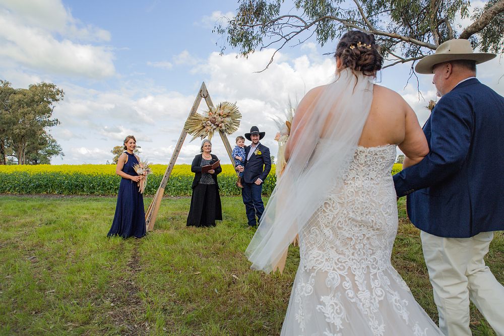 Wedding party gathered in a rural setting with bright yellow canola fields and eucalyptus trees in the background.