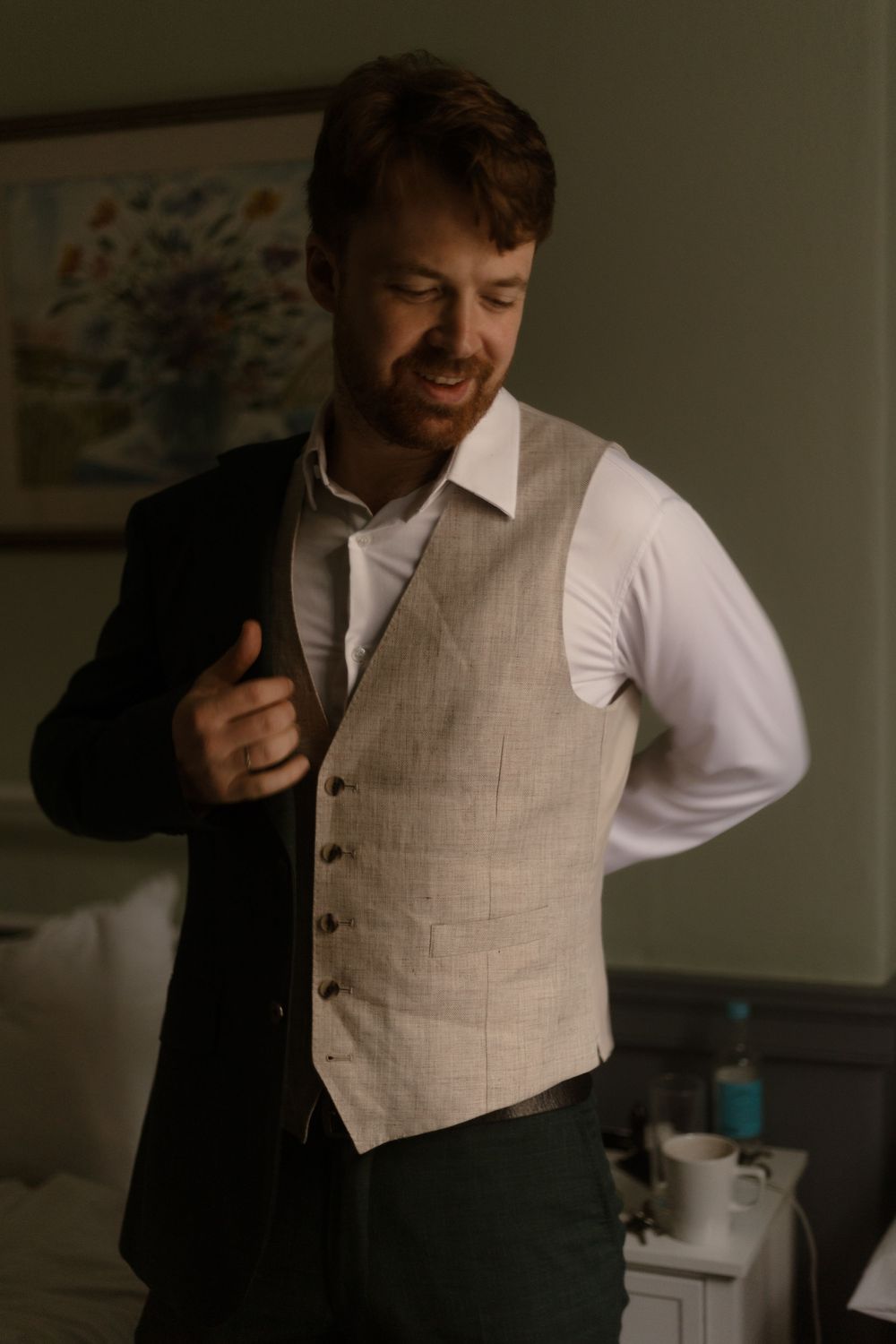 Person in a beige vest and white dress shirt adjusts their collar while holding a black coat in a dimly lit room.