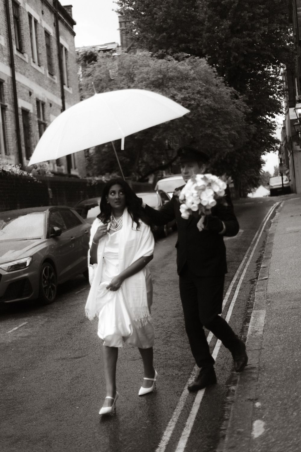 Wedding party walks down a rainy street with white umbrellas, creating a playful moment in black and white.