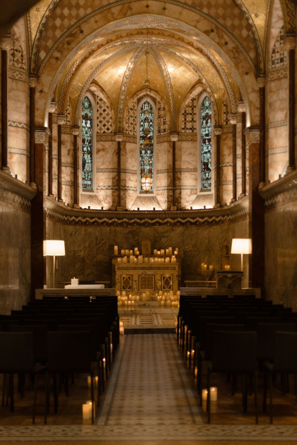 Gothic chapel interior with vaulted ceiling, stained glass windows and rows of dark wooden pews lit by soft lighting.