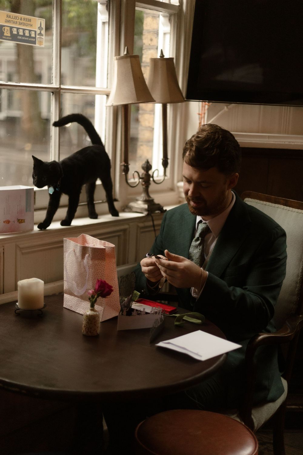 A black cat walks along a windowsill while someone sits at a wooden table working in moody lighting.