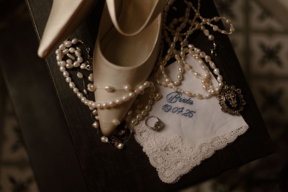 Elegant wedding accessories featuring white bridal shoes, pearls, and a delicate lace-trimmed note laid on dark fabric.