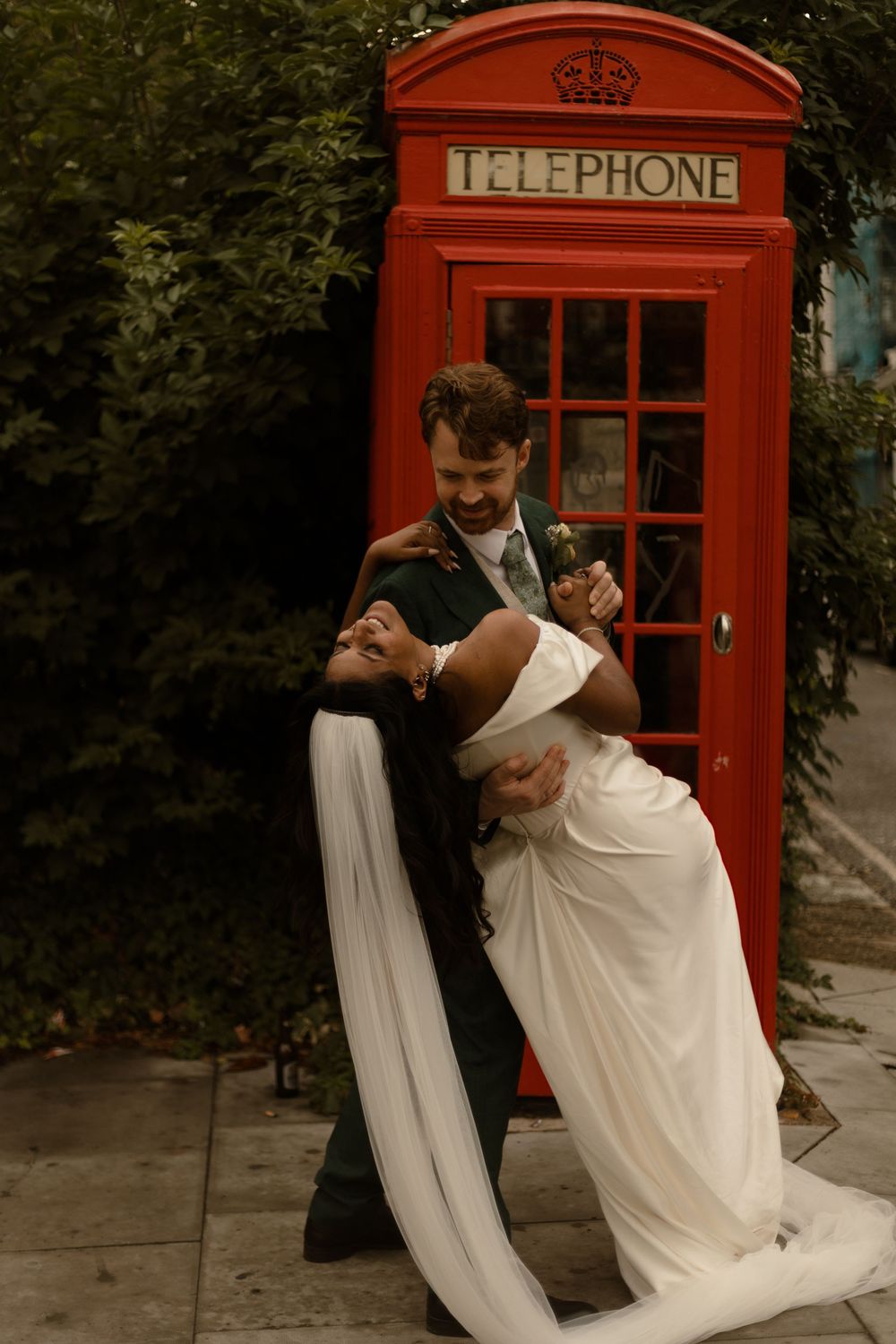 A romantic wedding couple poses in front of a classic red British telephone booth in an intimate dip pose.