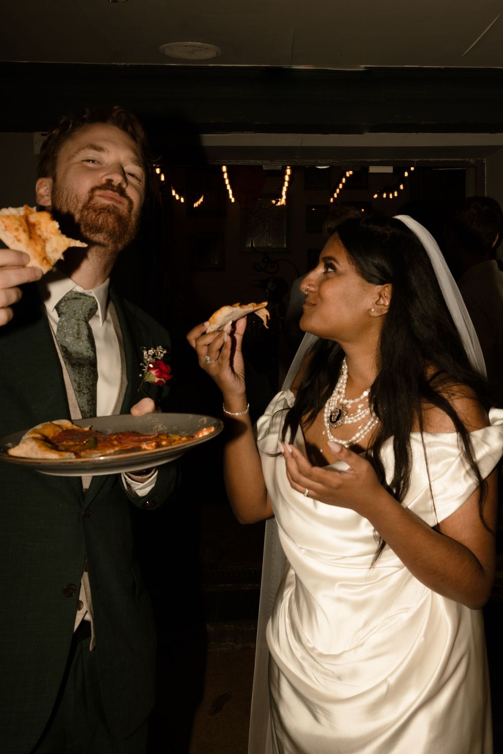 A couple eating cake together in formal wedding attire at a reception venue in low lighting.