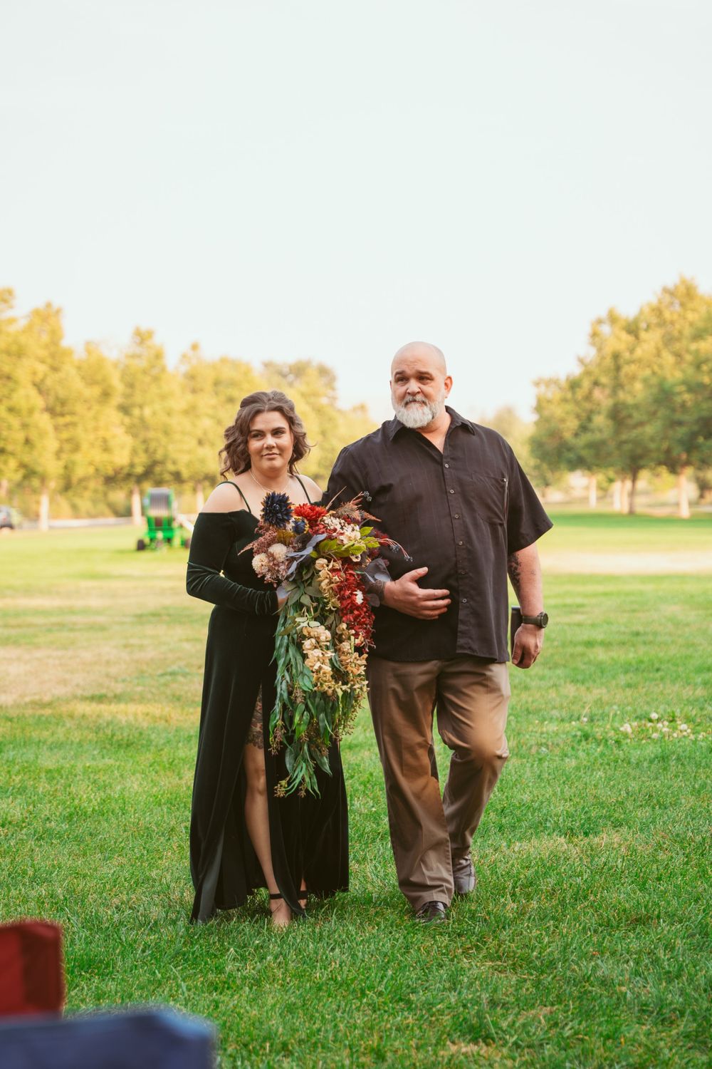 A couple stands together outdoors holding a vibrant red and purple floral bouquet.