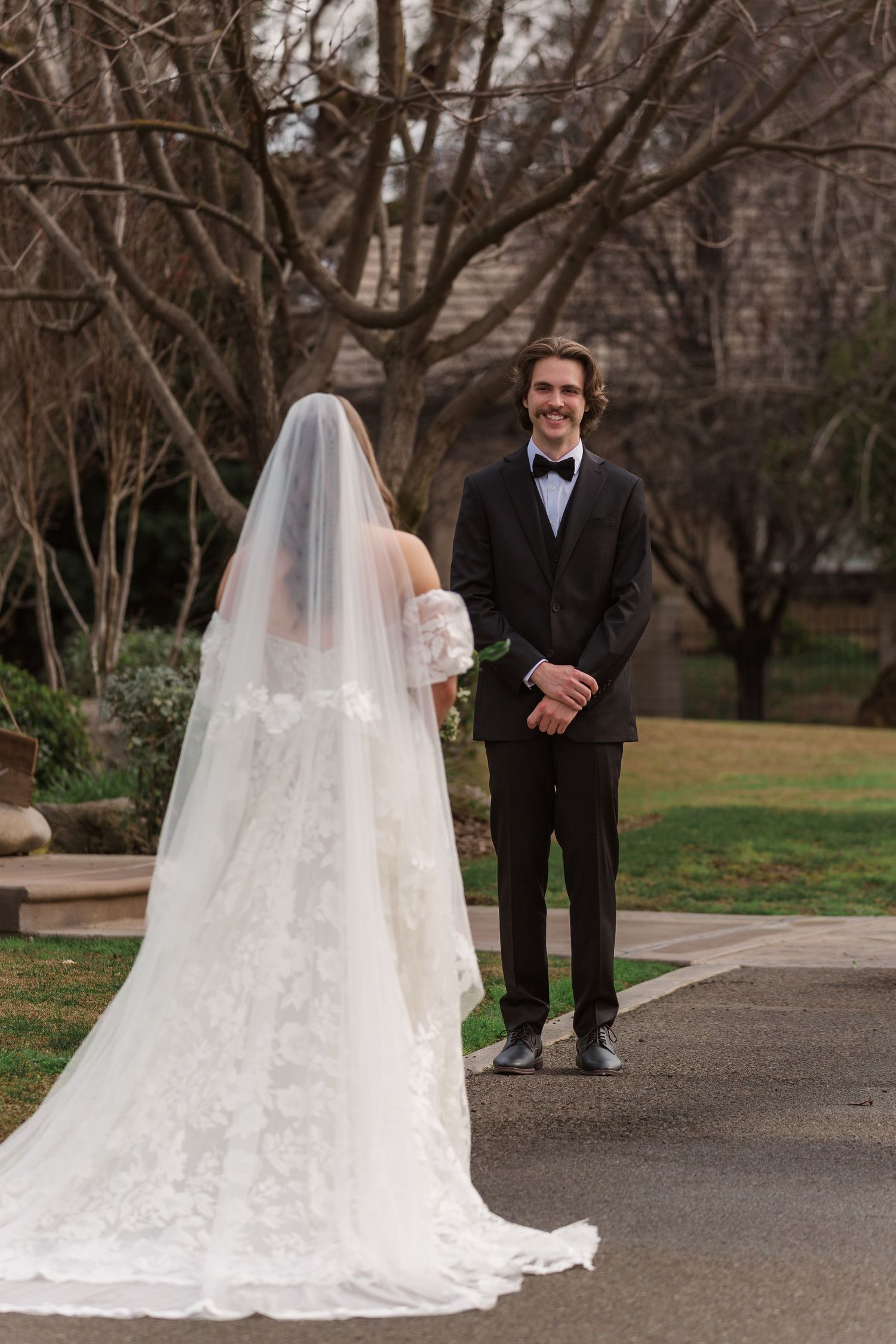 A bride in a flowing lace gown with long veil walks along a garden path.
