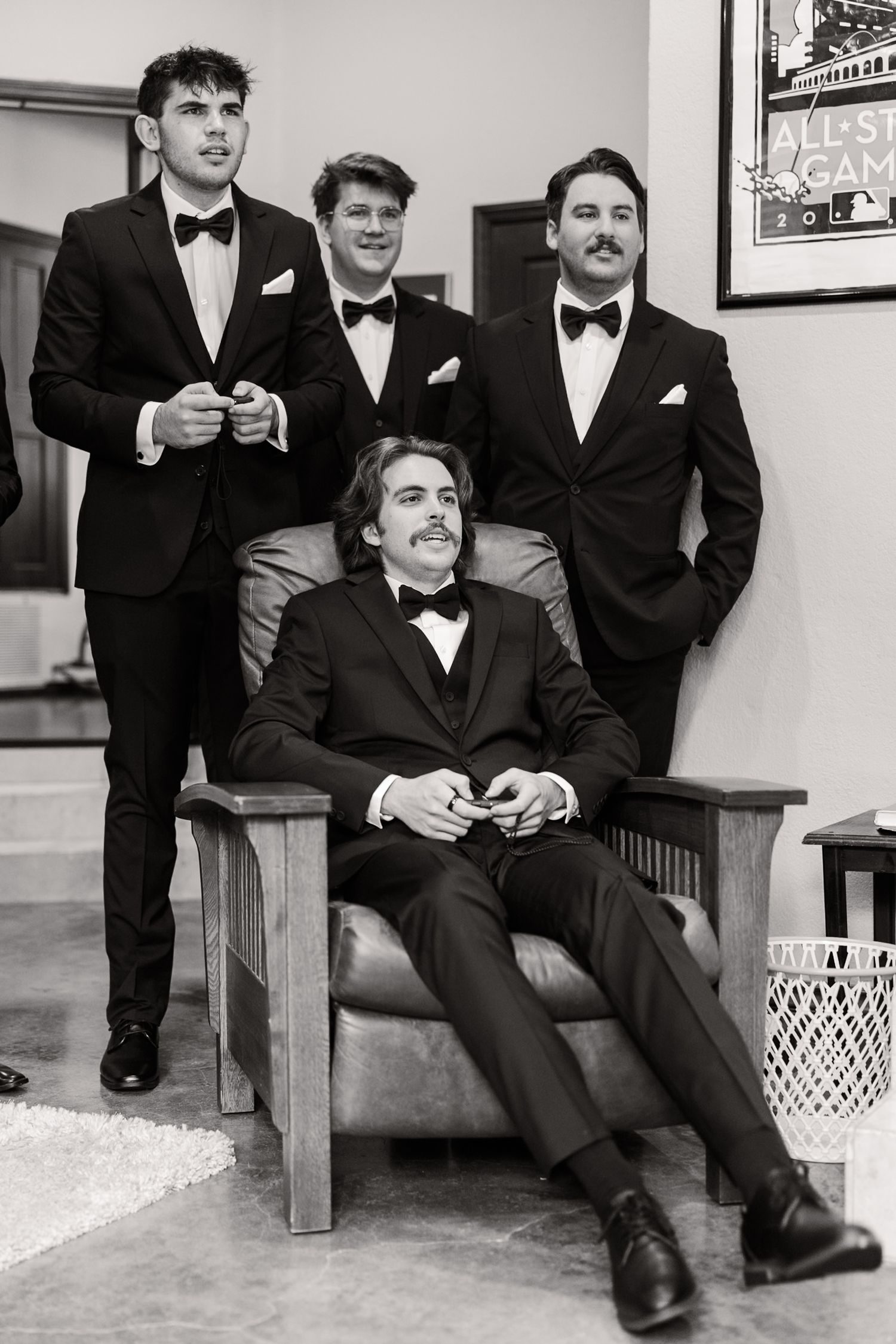 Groomsmen in black tuxedos relax on wooden benches in casual portrait series.