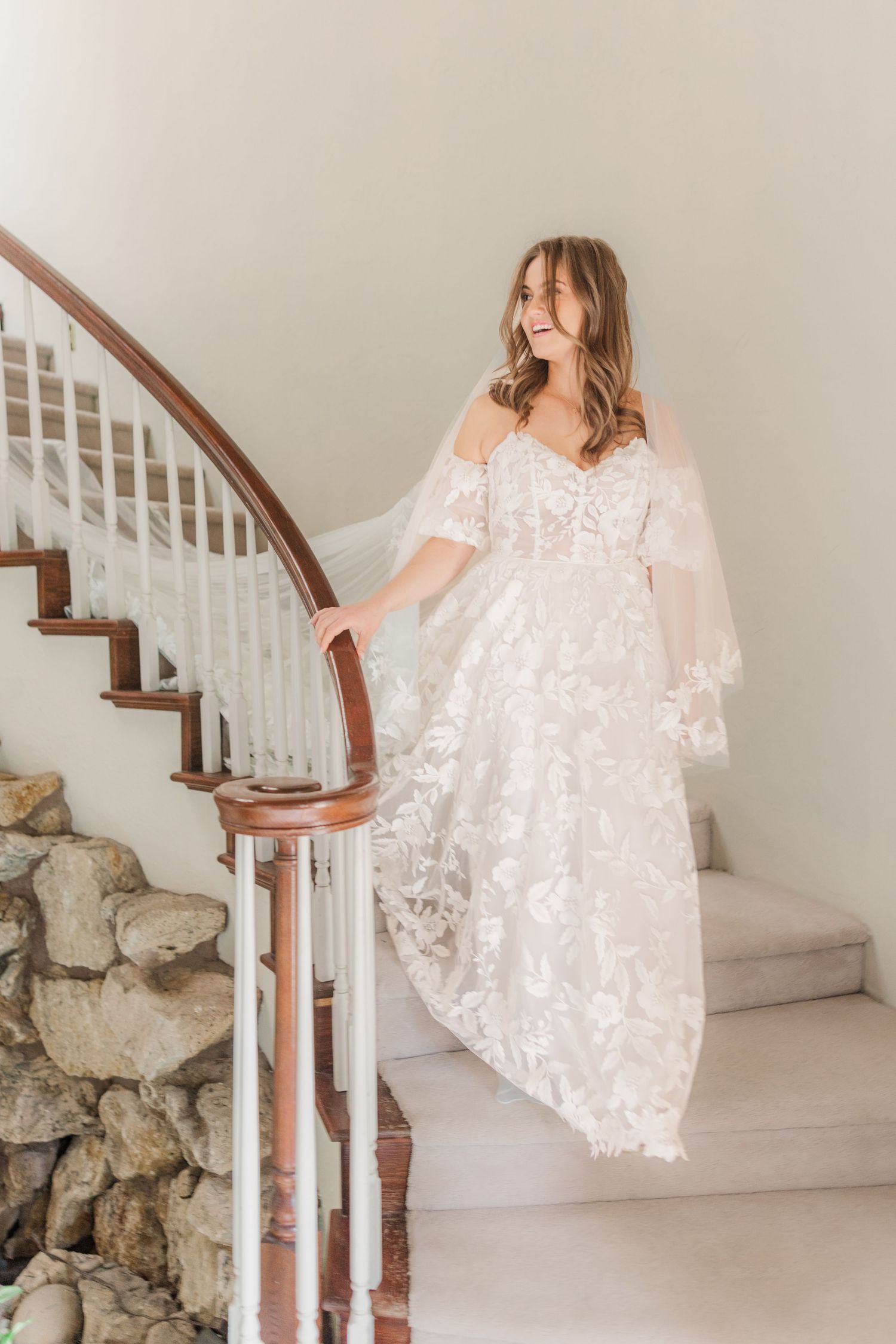 Bride in off-shoulder lace gown poses gracefully on white staircase.