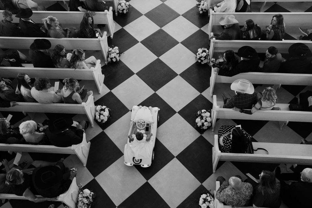 A black and white aerial view of people seated in church pews with a checkered floor pattern creating geometric shapes.
