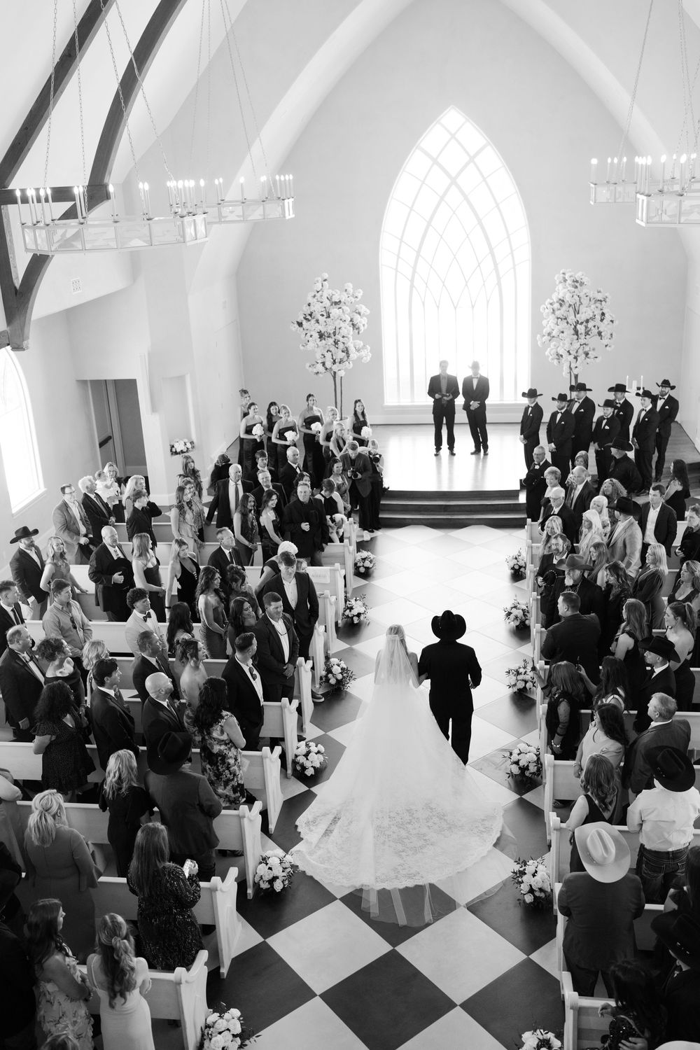 Aerial view of a wedding ceremony in a white chapel with gothic arched windows and black and white checkered floor.