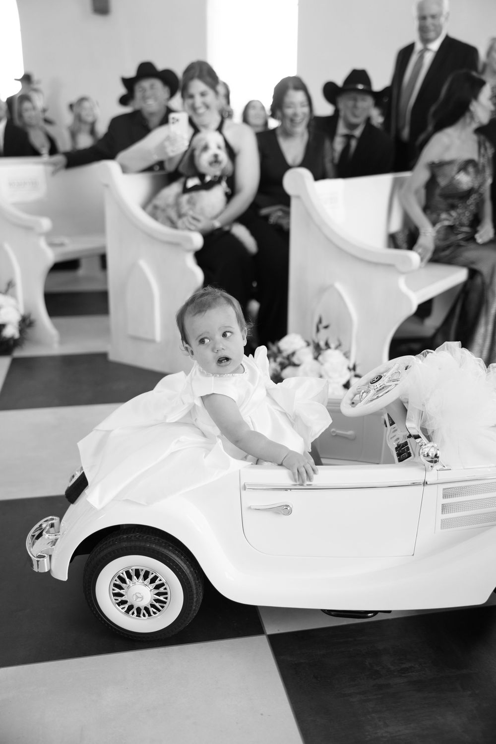 A young child in a white dress rides down the aisle in a miniature vintage-style white car during a wedding ceremony.
