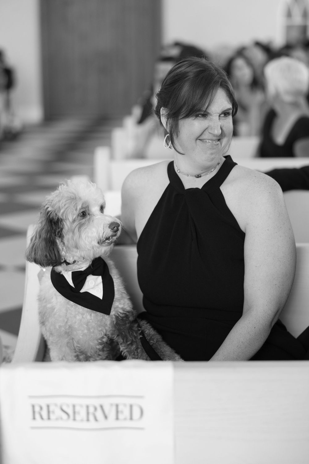 A person in a black halter dress sits with a small white dog wearing a bowtie at a reserved event seating area.