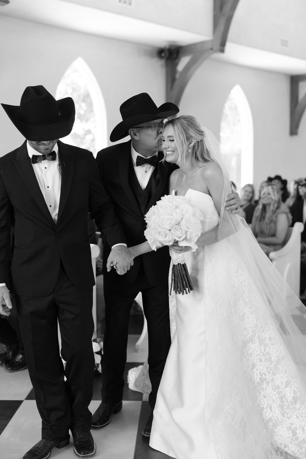 A black and white wedding photo in an elegant church setting with two men in cowboy hats and a bride in white gown.