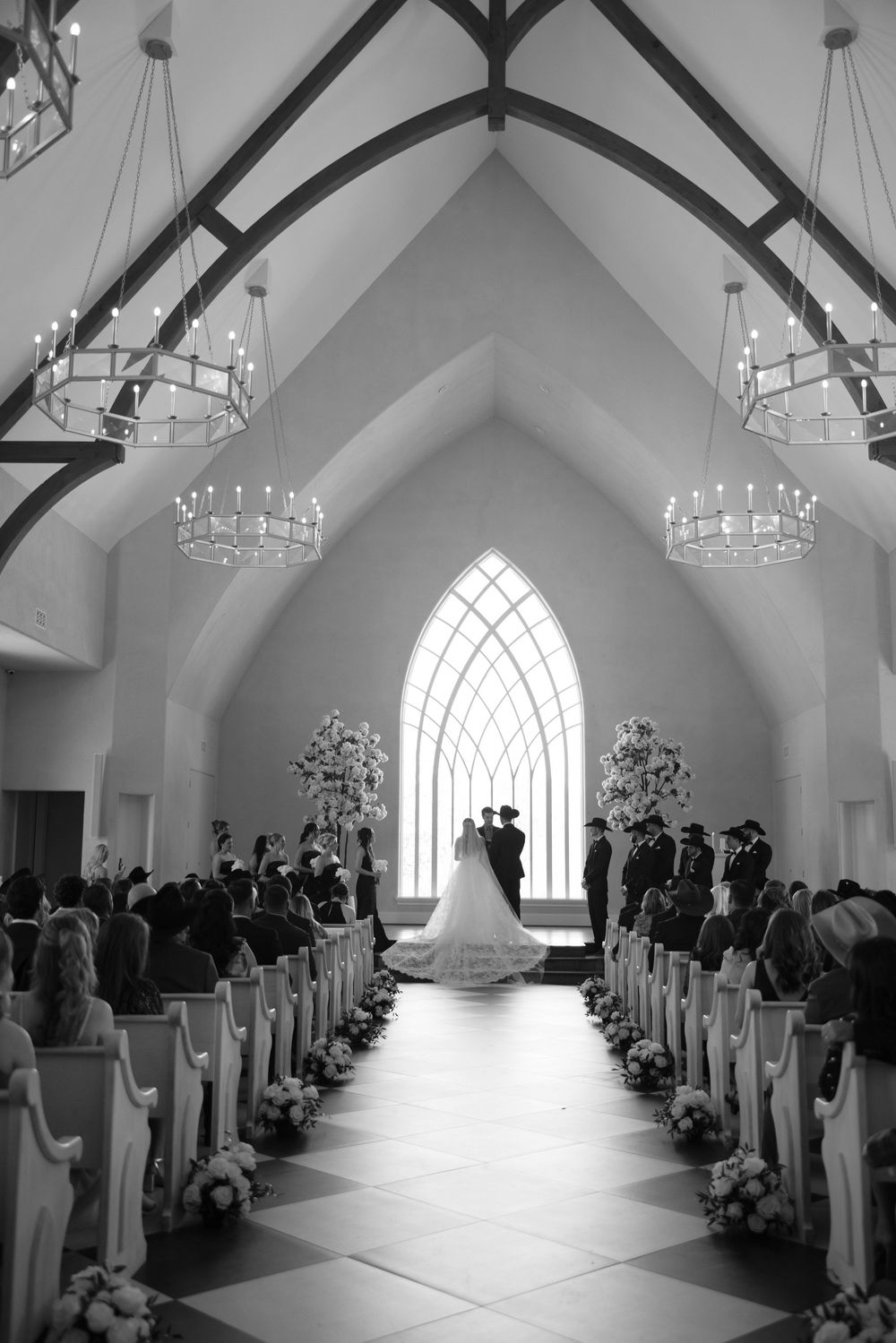Black and white photograph of a wedding ceremony in a chapel with soaring arched ceiling, chandeliers and a gothic window.