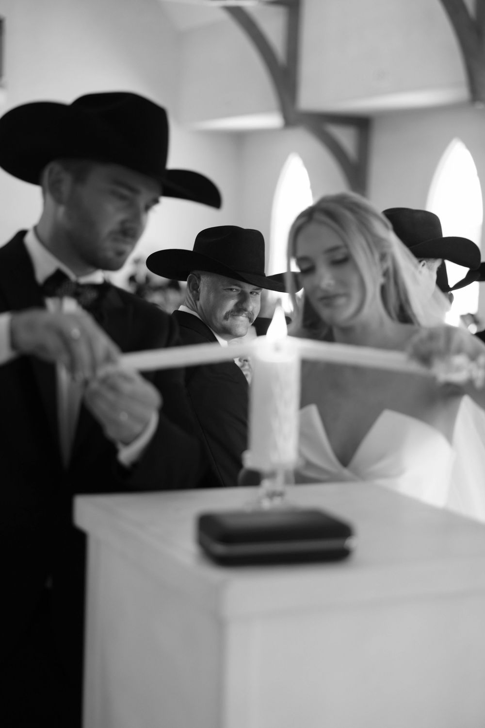 A bride in white and groomsmen in black cowboy hats participate in a unity candle ceremony at a chapel wedding.