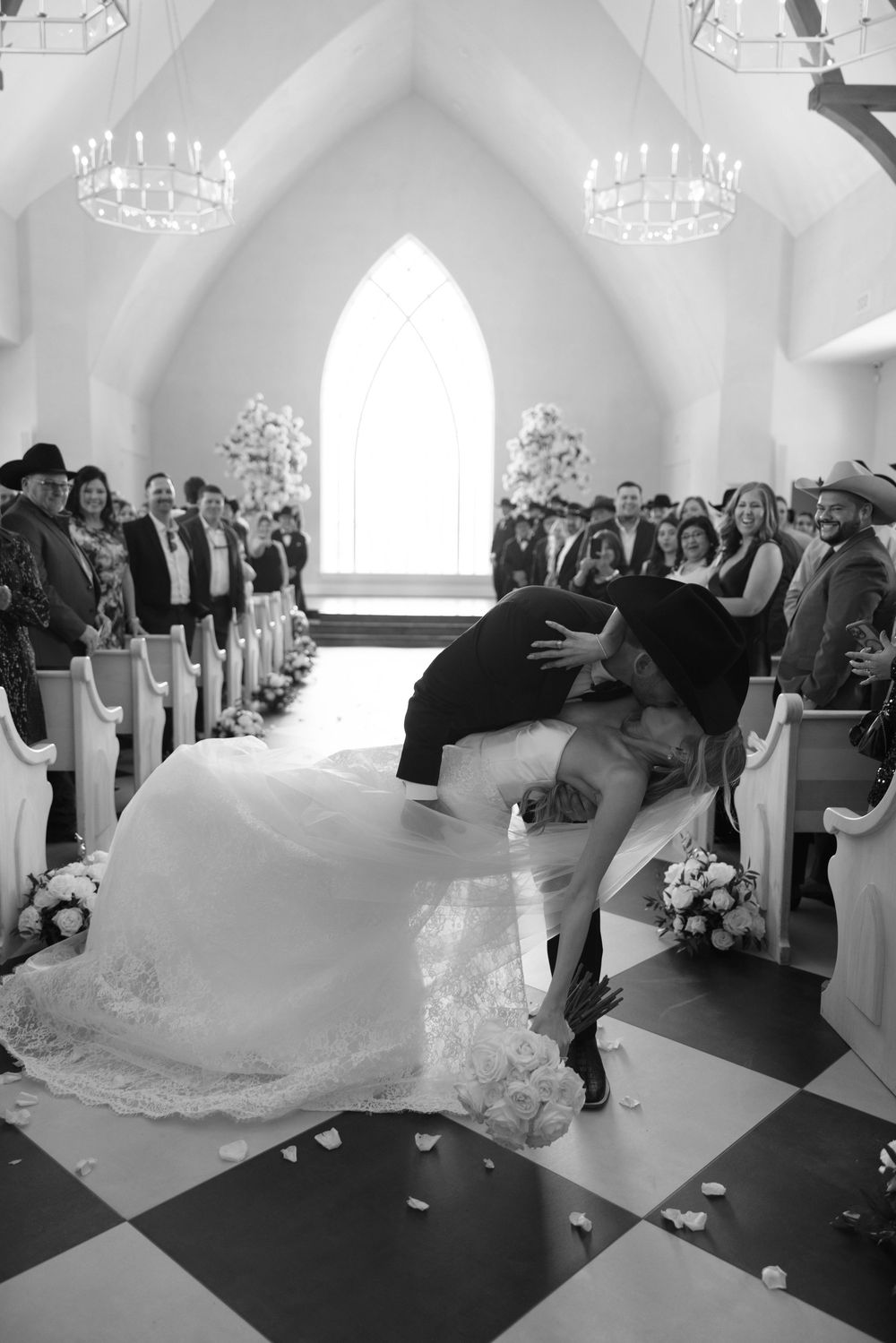Black and white photo of a dramatic dip kiss in a chapel aisle with chandeliers and guests watching during a wedding ceremony.