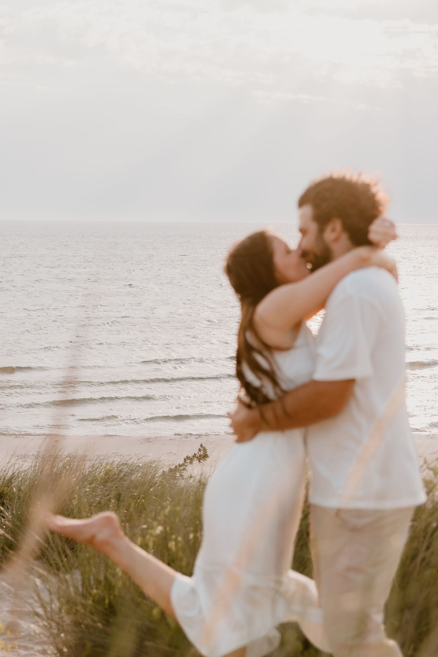 A romantic couple embraces at sunset on a beach with tall beach grass in the foreground. Metro Detroit Engagement and Wedding Photographer