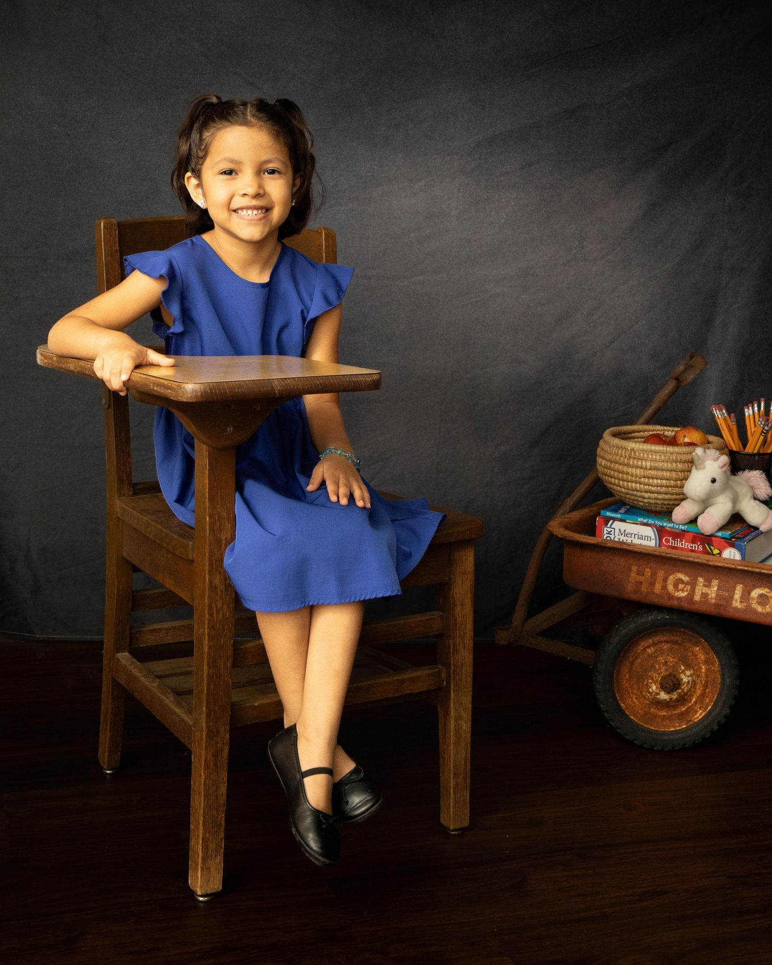 A child in a blue dress sits on a wooden school chair against a dark background.